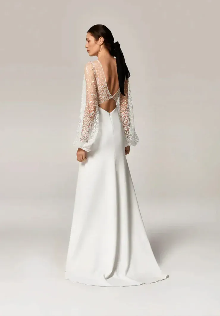 A woman modeling a white wedding gown from the back, showing a long open back, sheer lace long sleeves, and a floor-length satin skirt against a plain light gray studio background.