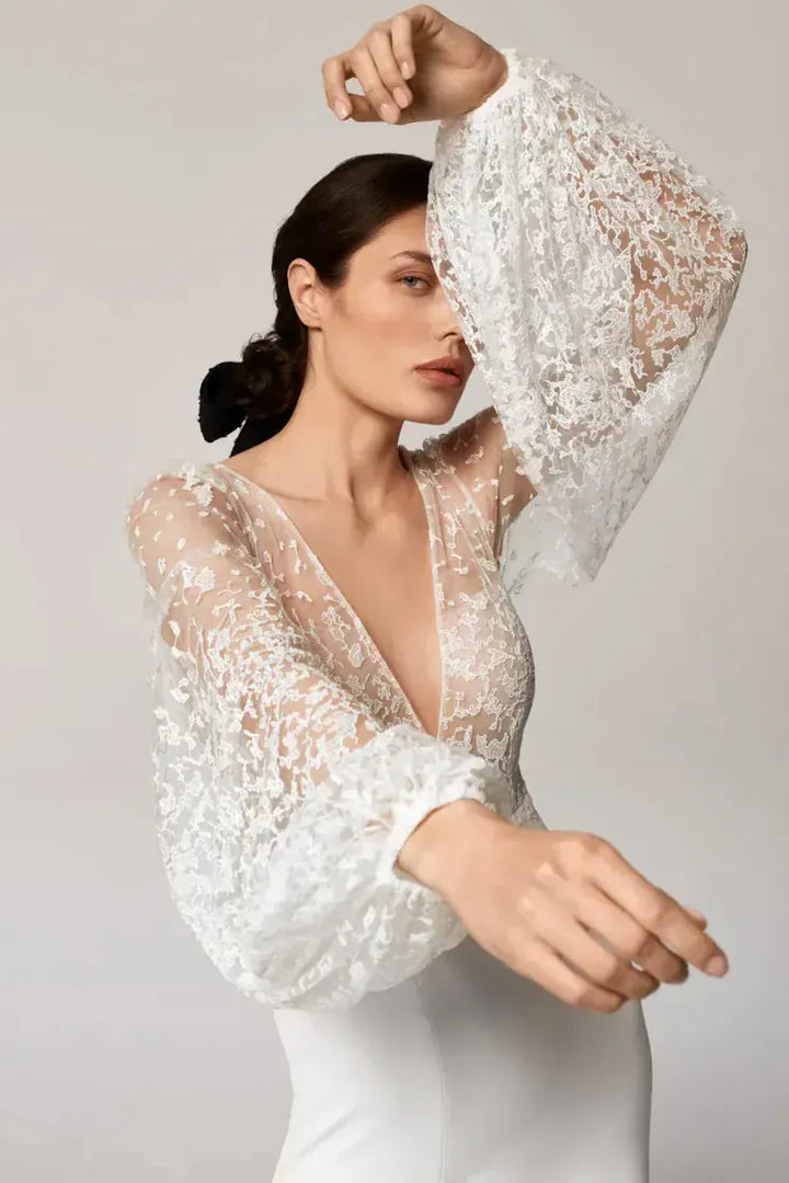 A woman modeling a white lace long-sleeve dress with a deep V-neckline and sheer balloon sleeves, posed against a light gray studio background.