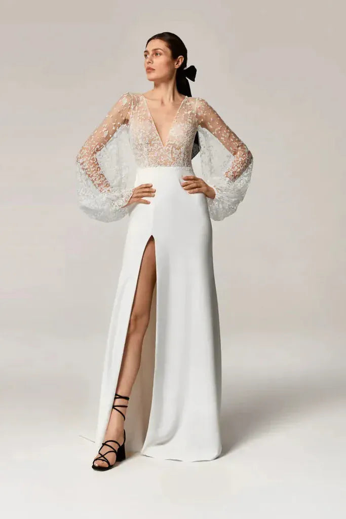 A woman models a floor-length white gown with a high thigh slit, a deep V-neckline, and sheer lace sleeves with balloon sleeves, posed against a neutral light beige studio background.