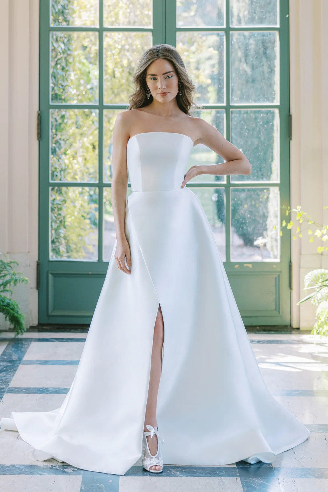 Woman in a white strapless gown with a high slit standing in front of large windows.