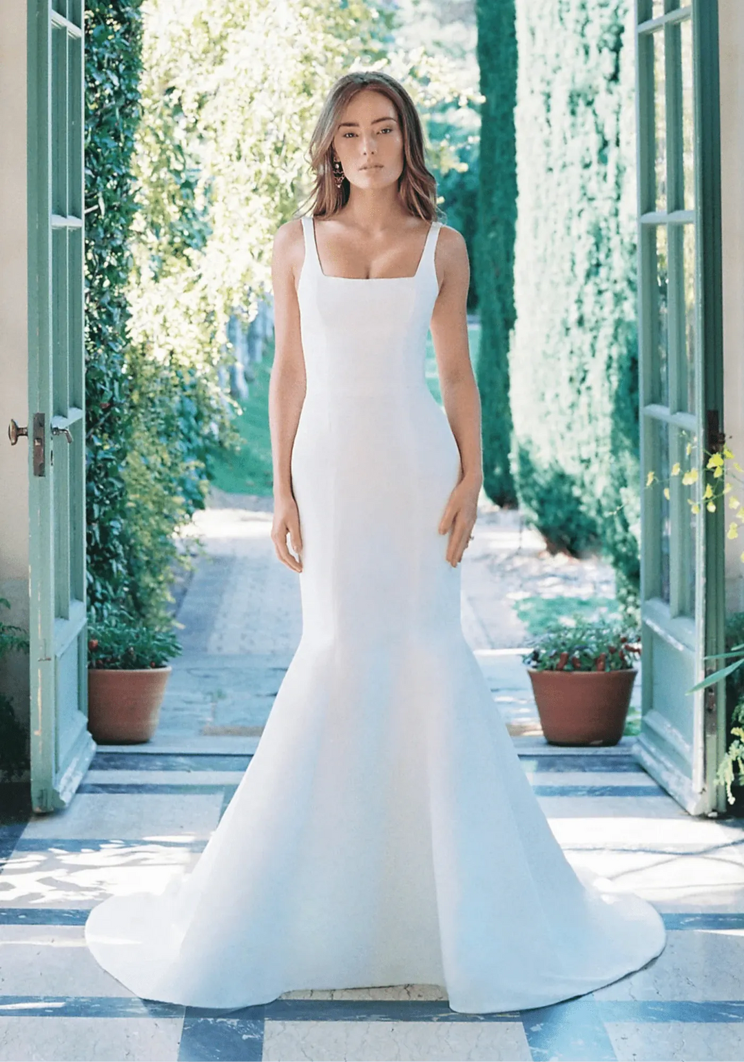 Woman in a white wedding dress standing in a sunlit outdoor setting with greenery.