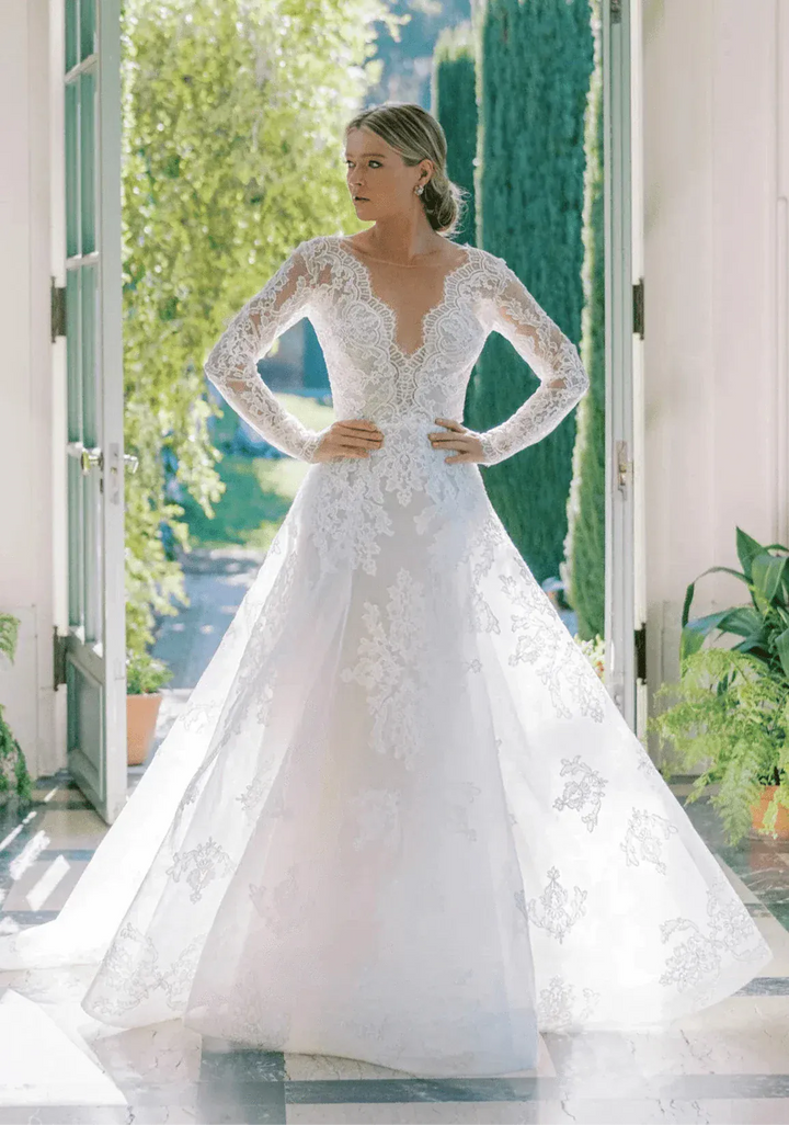 Woman wearing a white lace wedding dress in a bright room with greenery outside.