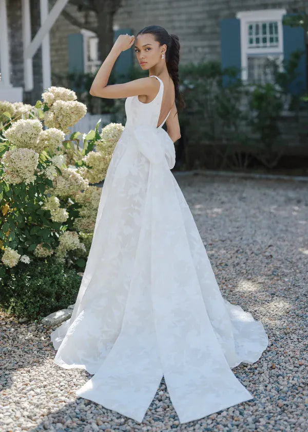 Woman in a white wedding dress standing outdoors with flowers and a building in the background