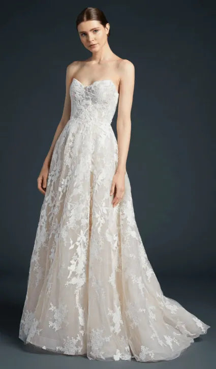 A woman modeling a strapless A-line wedding gown with a sweetheart neckline, fitted lace bodice, and a full tulle skirt with floral lace overlay, photographed against a dark gray studio background.