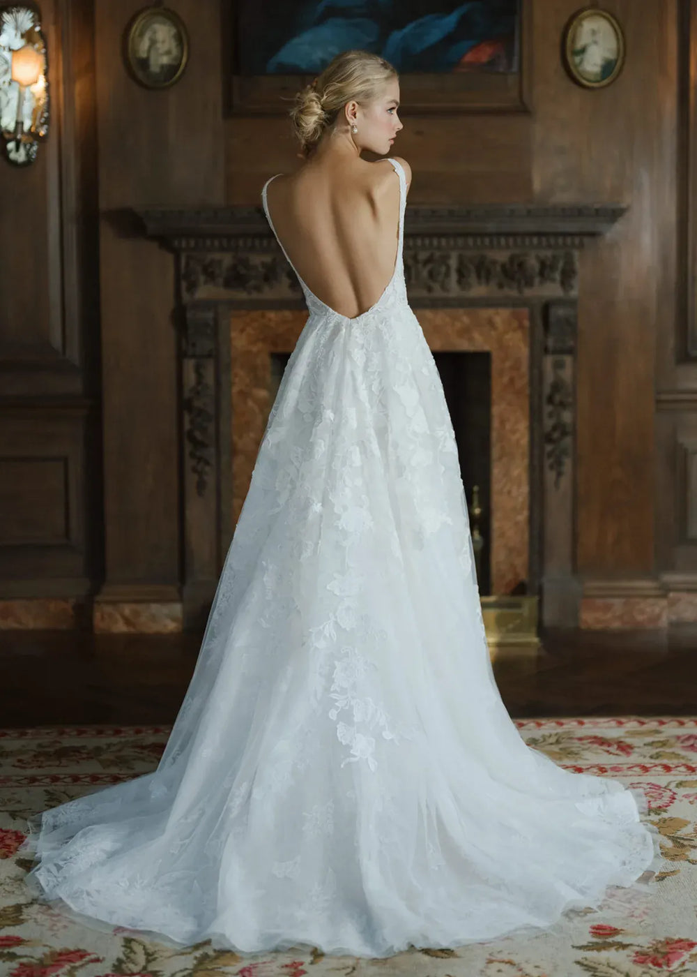 Woman in a white wedding dress with an open back standing in a room with wooden paneling and a fireplace.