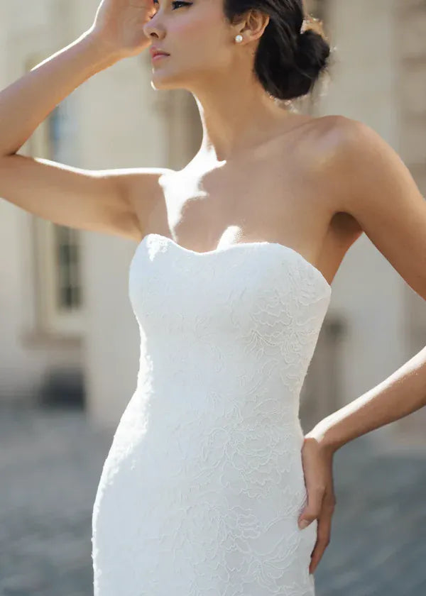 A woman wearing a strapless white lace wedding gown, photographed outdoors with a softly blurred light-colored building in the background.