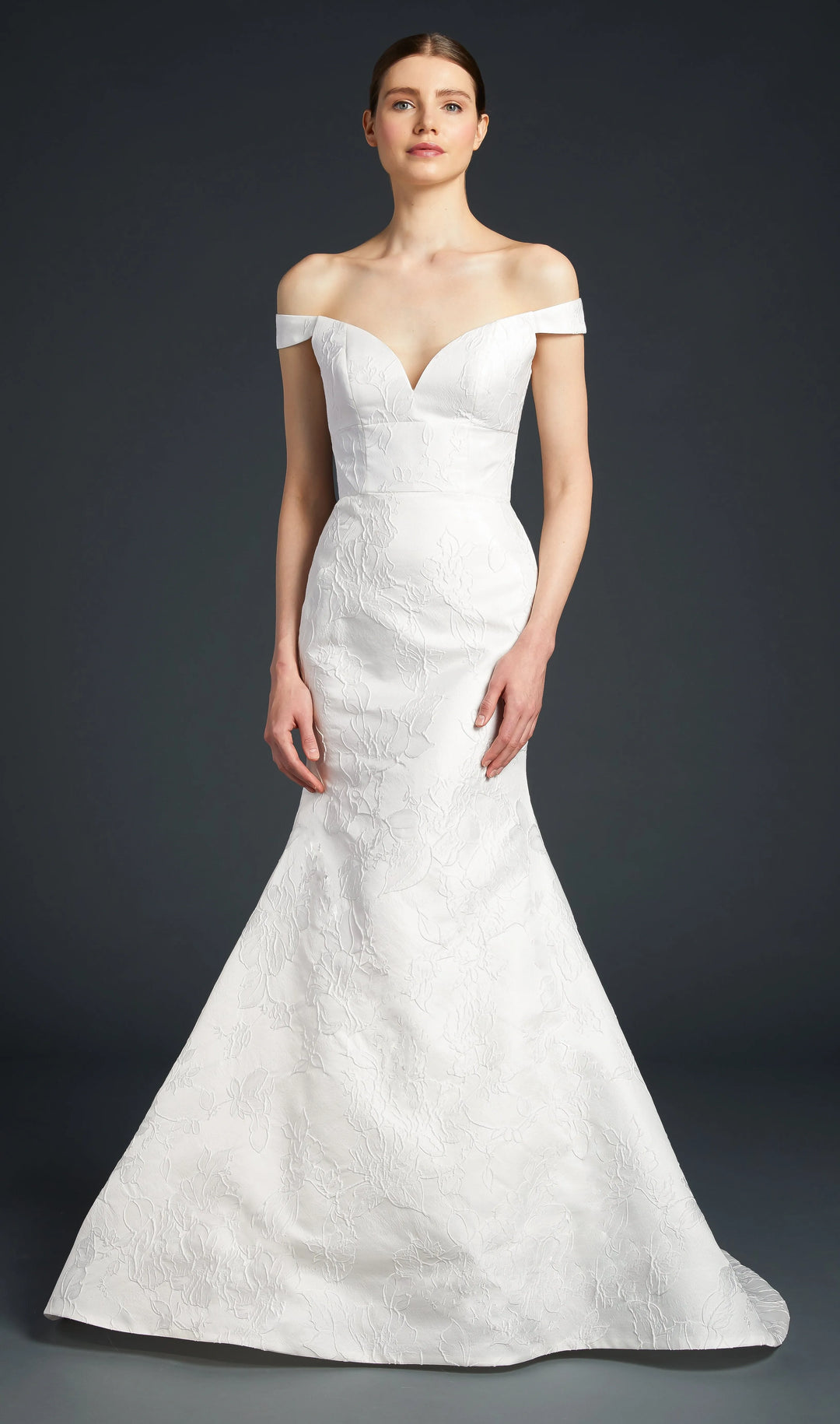 A woman modeling an off-the-shoulder white wedding gown with a fitted bodice and flared mermaid skirt against a dark gray studio background.