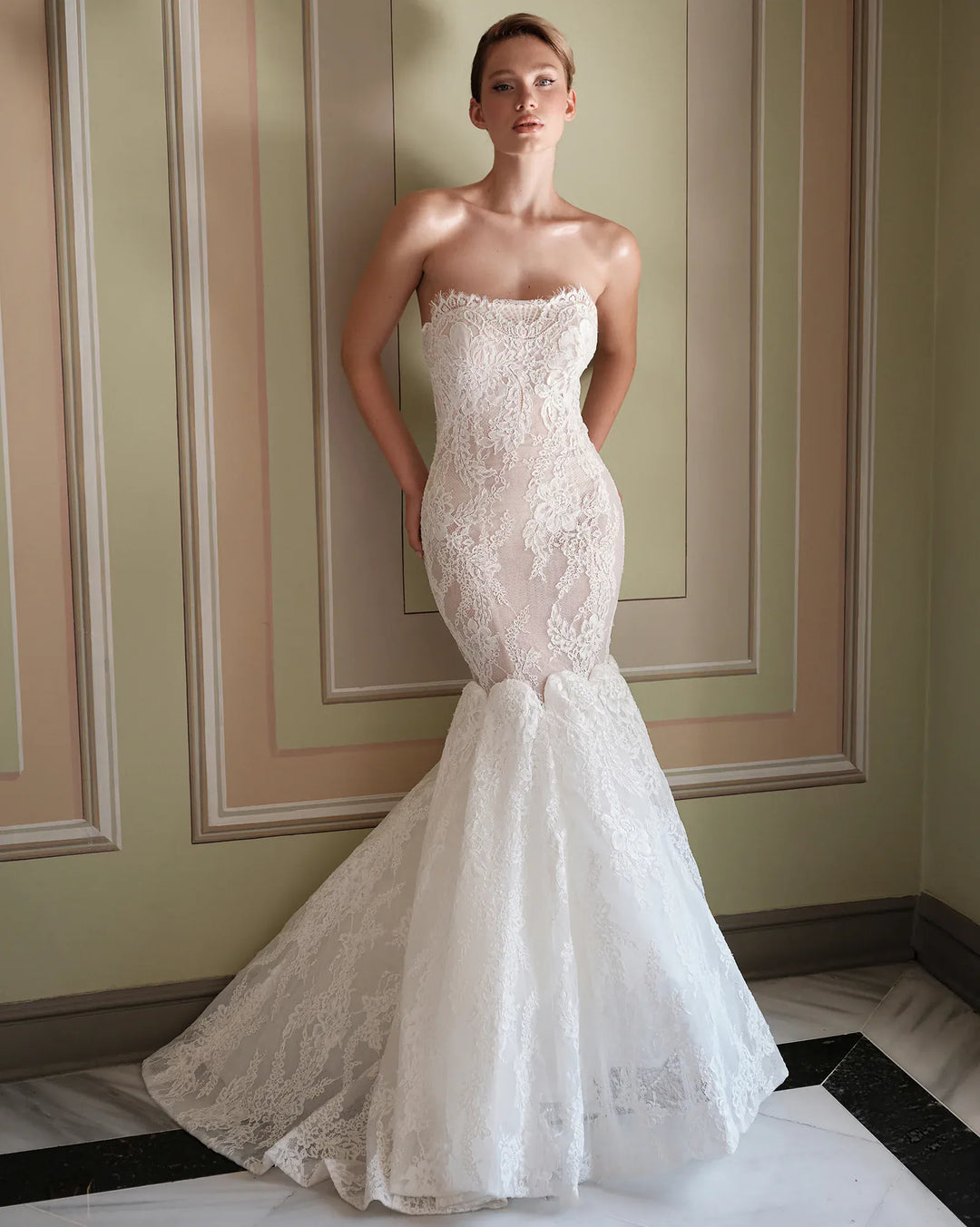A woman models a strapless white lace mermaid wedding gown in an elegant indoor setting with paneled walls and a marble floor.