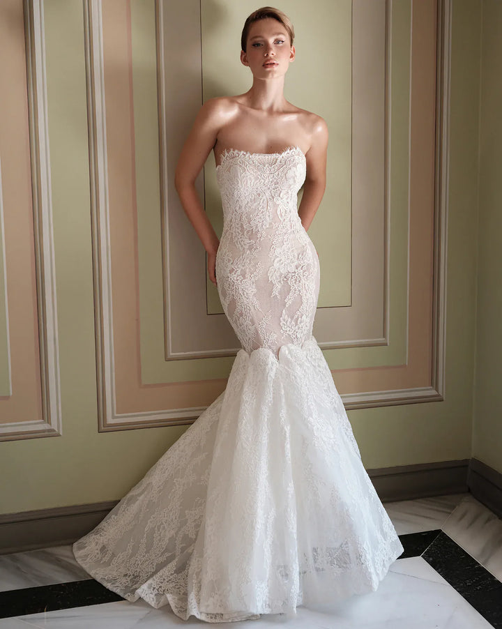A woman models a strapless white lace mermaid wedding gown in an elegant indoor setting with paneled walls and a marble floor.