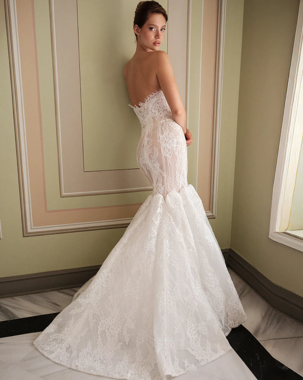 A woman modeling a strapless white lace mermaid wedding gown, photographed indoors against a paneled wall with a window light on the right.