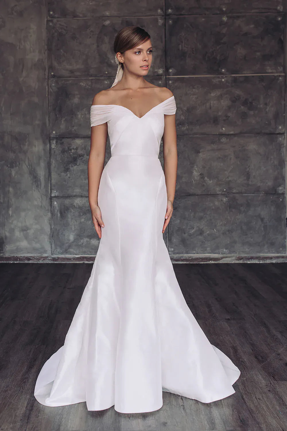 Woman wearing a white off-shoulder wedding dress against a dark textured wall.