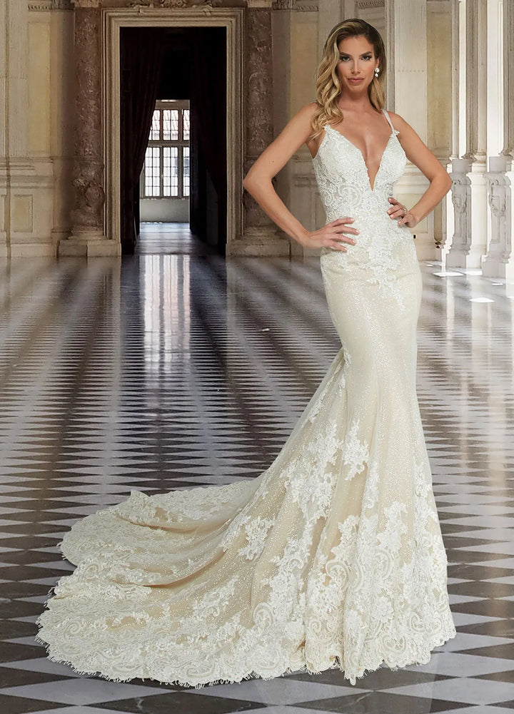 A woman modeling a white lace mermaid-style wedding gown with a deep V-neckline and long train in an elegant indoor hall with a checkered floor.