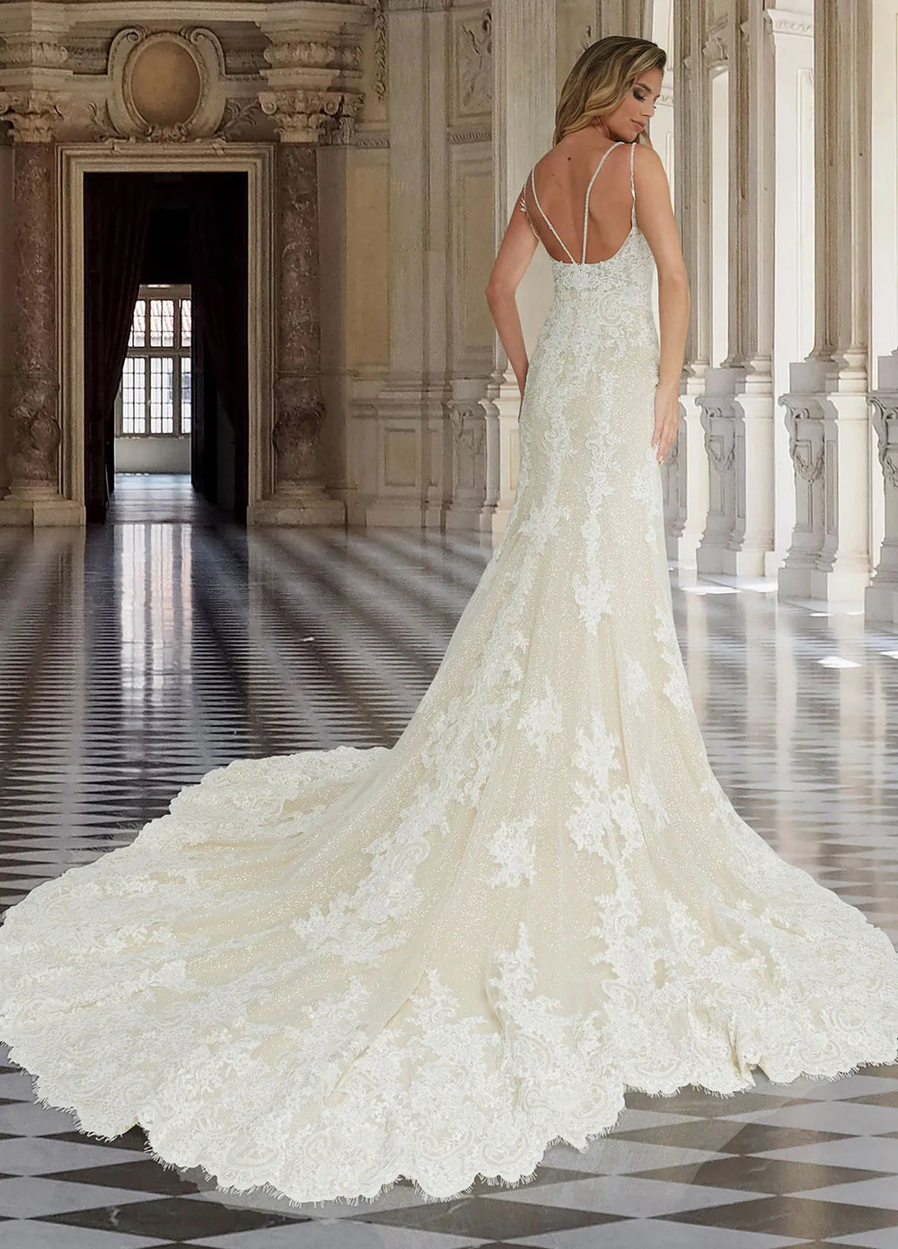 A woman wearing a sleeveless ivory lace wedding gown with thin straps and a dramatic train, photographed from the back in a grand classical hall with columns and a tiled floor.