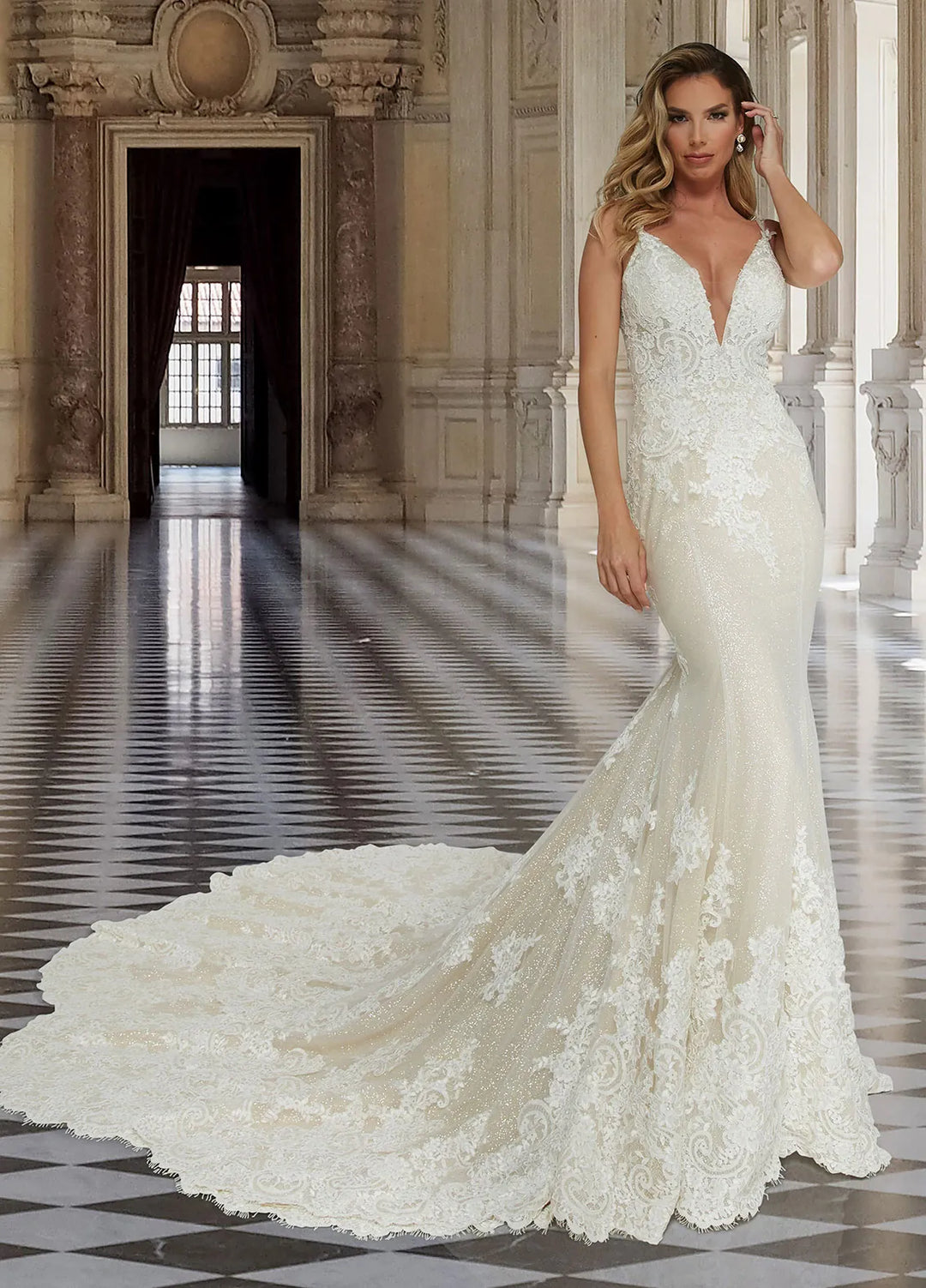 A woman modeling a white lace mermaid-style wedding gown with a deep V neckline, fitted silhouette, and long train in an ornate indoor hall with a checkered floor.