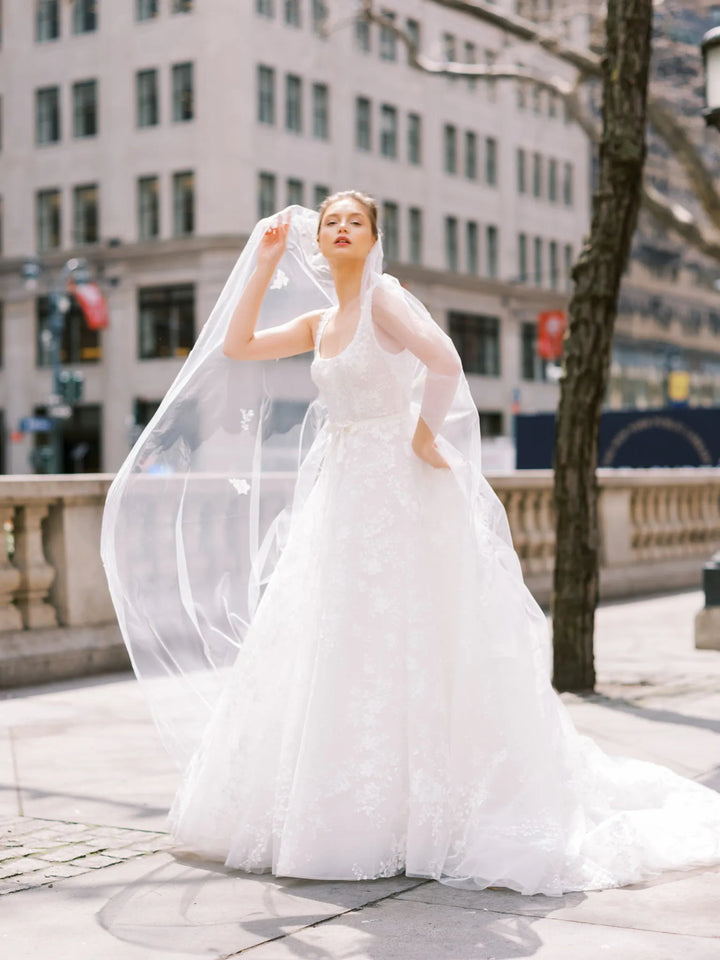 A woman wearing a white lace wedding gown with a long sheer veil stands outdoors on a city sidewalk in front of a building and tree.