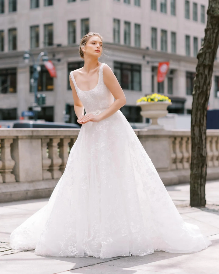 A woman wearing a white lace wedding gown stands outdoors on a city sidewalk in front of a large stone building, with a tree trunk visible on the right.