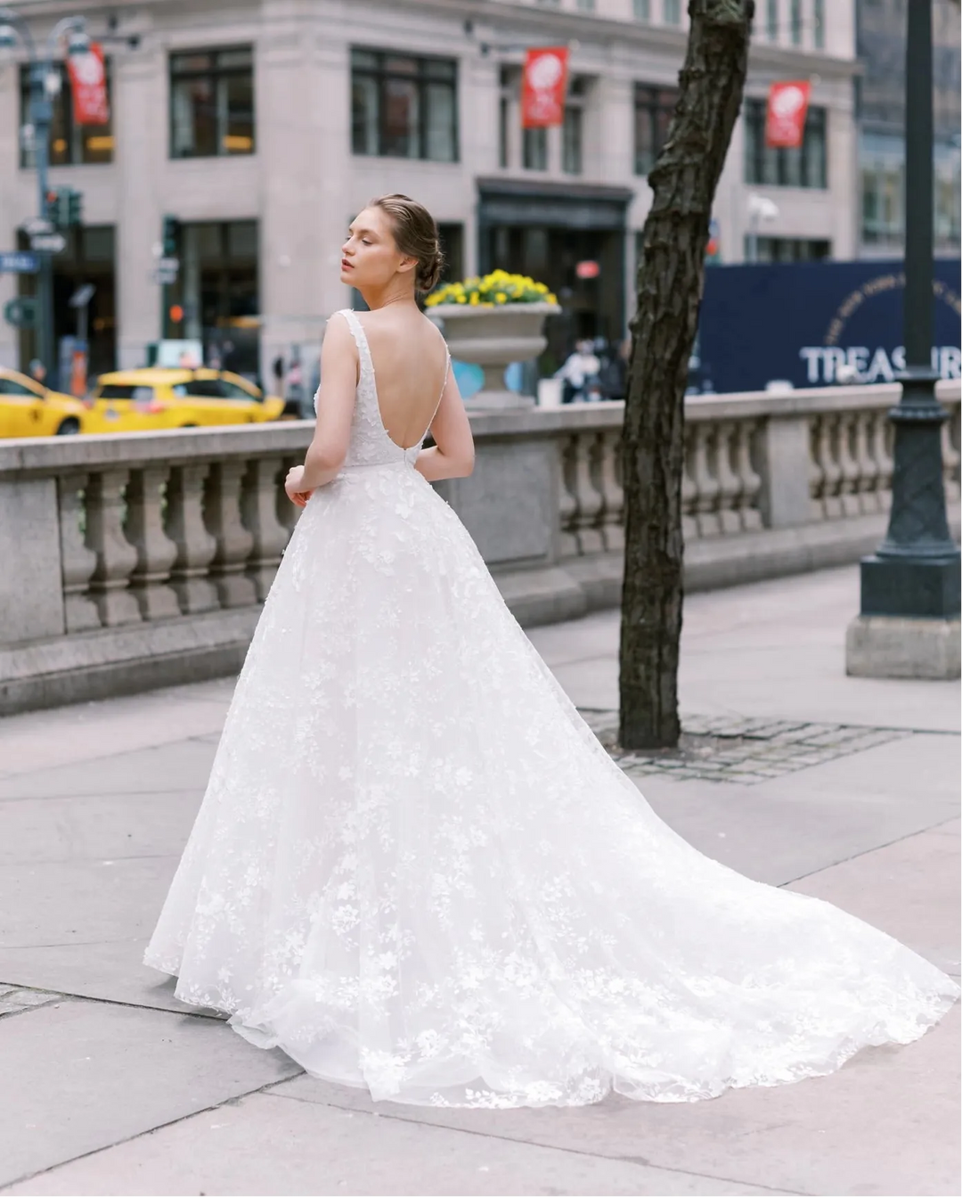 A woman wearing a white lace wedding gown stands on a city sidewalk, photographed from behind as she looks upward. The dress has a fitted bodice, sheer straps, a low open back, and a full flared skirt with a long train.
