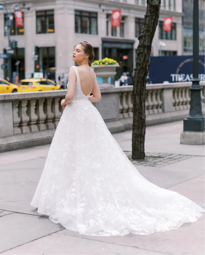 A woman wearing a white lace wedding gown stands on a city sidewalk, photographed from behind as she looks upward. The dress has a fitted bodice, sheer straps, a low open back, and a full flared skirt with a long train.