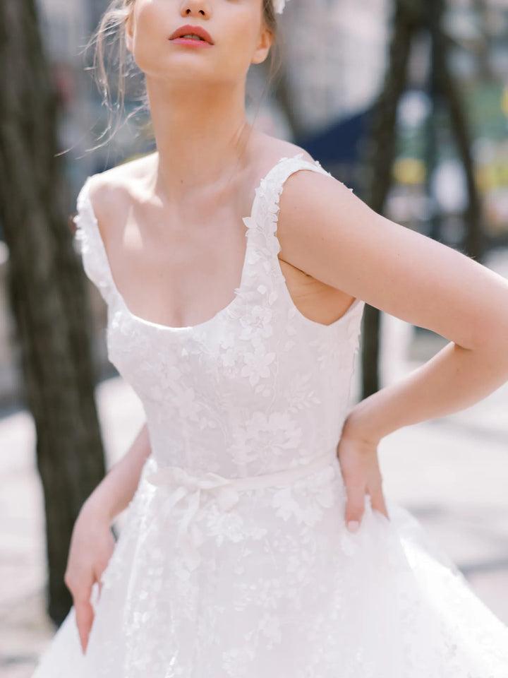 A woman wearing a white sleeveless lace wedding dress with a scoop neckline and a full skirt, photographed outdoors with blurred trees in the background.