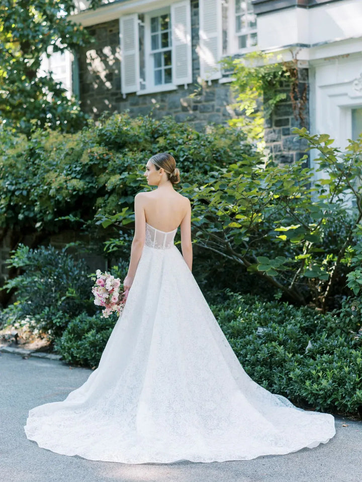 A bride in a white lace wedding gown stands outdoors in front of a stone house and greenery, photographed from behind to show the back of the dress with a long train and a small bouquet in hand.