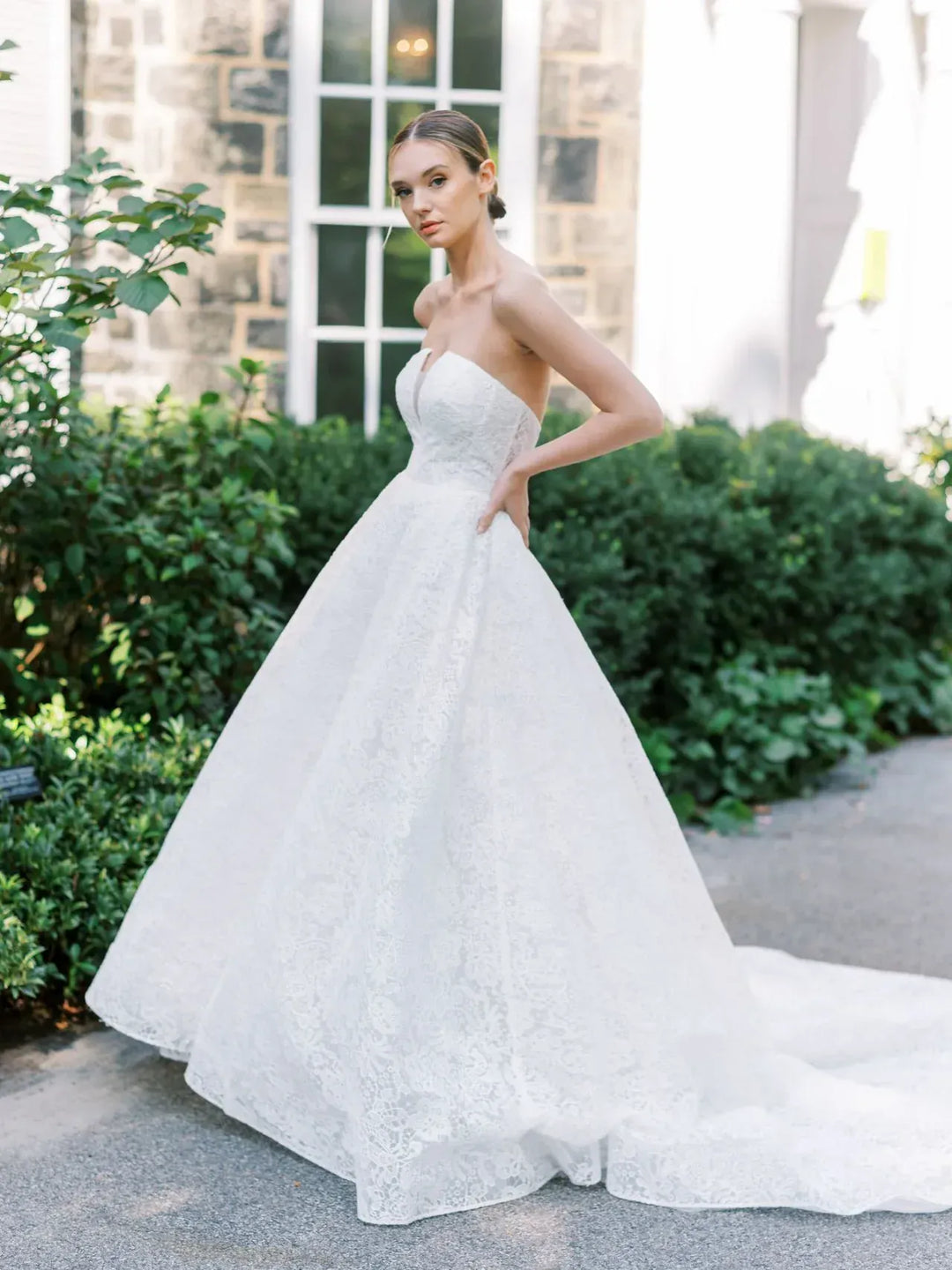 A woman wearing a strapless white lace ball gown wedding dress stands outdoors on a paved walkway with green shrubs and a stone building in the background.