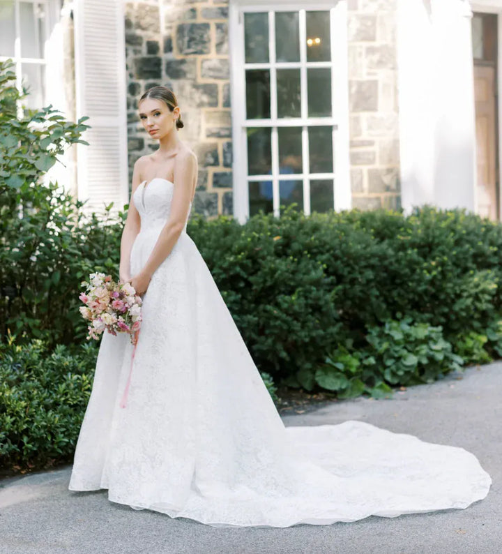 A woman wearing a strapless white lace wedding gown with a long train stands outdoors in front of a stone building, holding a bouquet of pink and cream flowers.