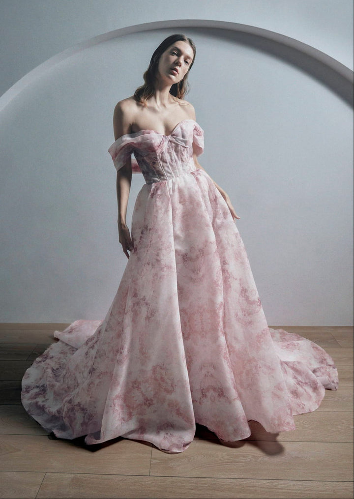A woman models an off-the-shoulder pink floral ball gown with a structured bodice and a voluminous skirt, posed in front of a minimalist gray background with a curved architectural element.