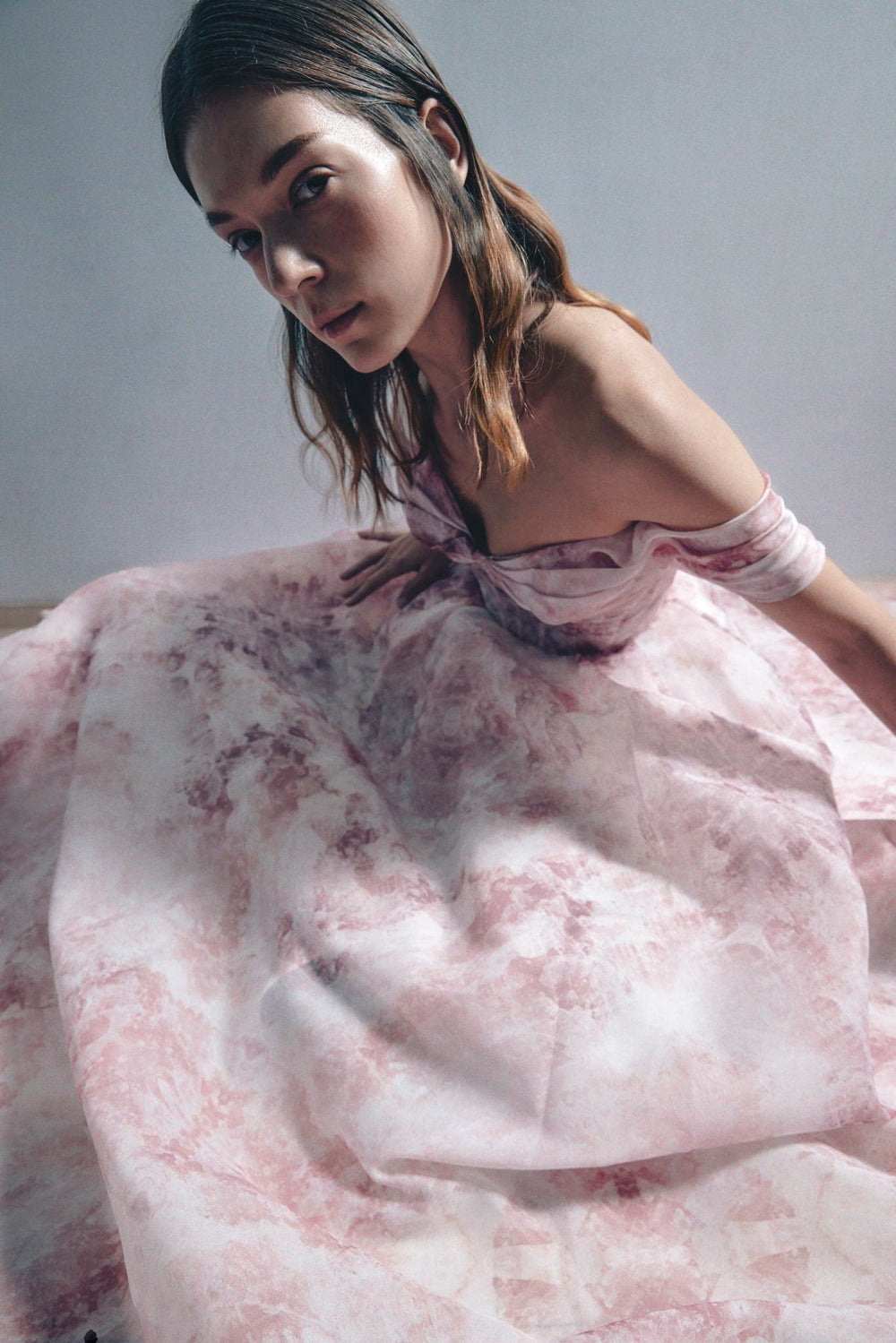 A model wearing a sheer, off-the-shoulder pink and white patterned dress or fabric drape, posed against a neutral gray background.