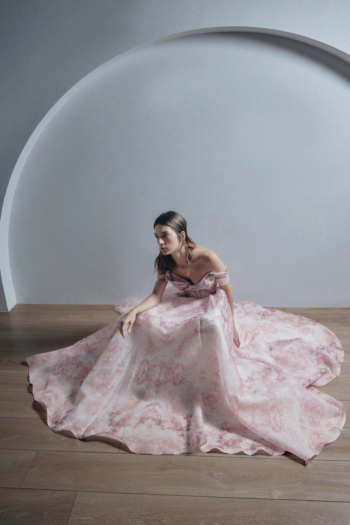 A woman wearing a voluminous off-the-shoulder pink gown with a floral jacquard pattern, sitting on a wooden floor in front of a large white semi-circular architectural backdrop.