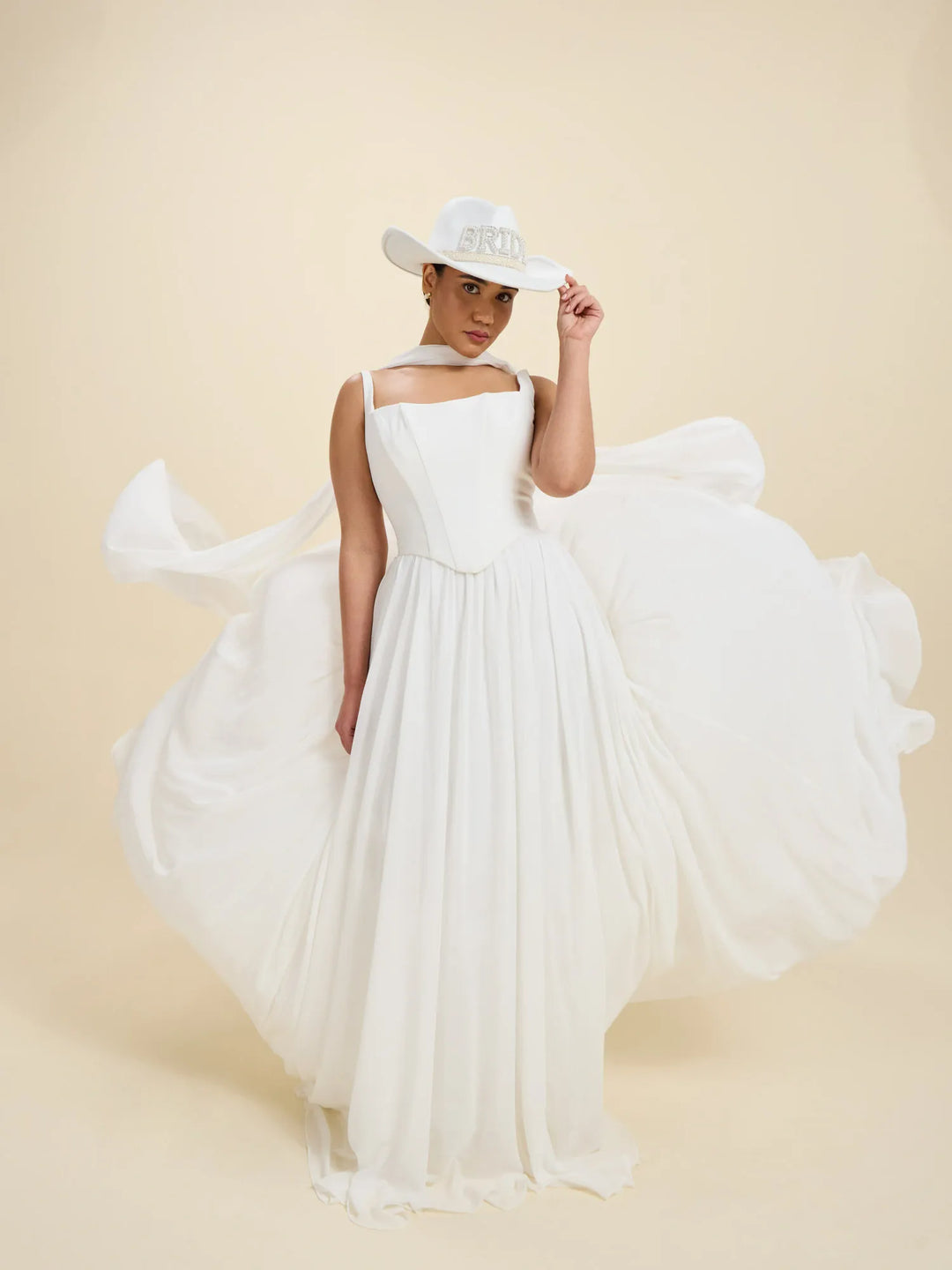 A woman wearing a flowing white wedding dress with a structured bodice and a large overskirt, posed in a studio against a warm beige background. She is also wearing a white cowboy hat with a silver band.