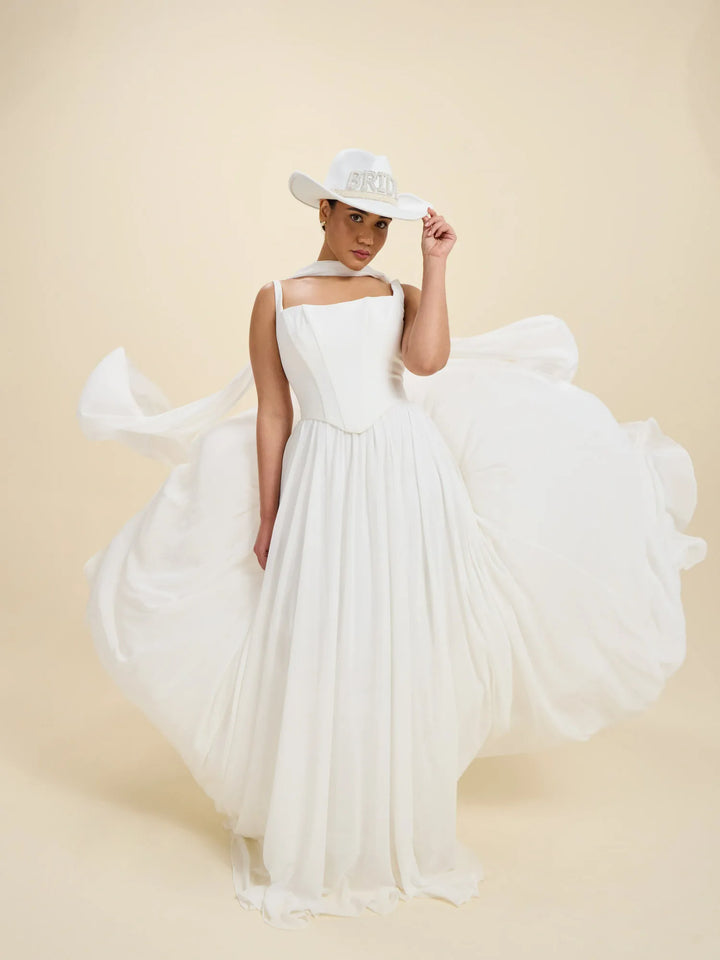 A woman wearing a flowing white wedding dress with a structured bodice and a large overskirt, posed in a studio against a warm beige background. She is also wearing a white cowboy hat with a silver band.