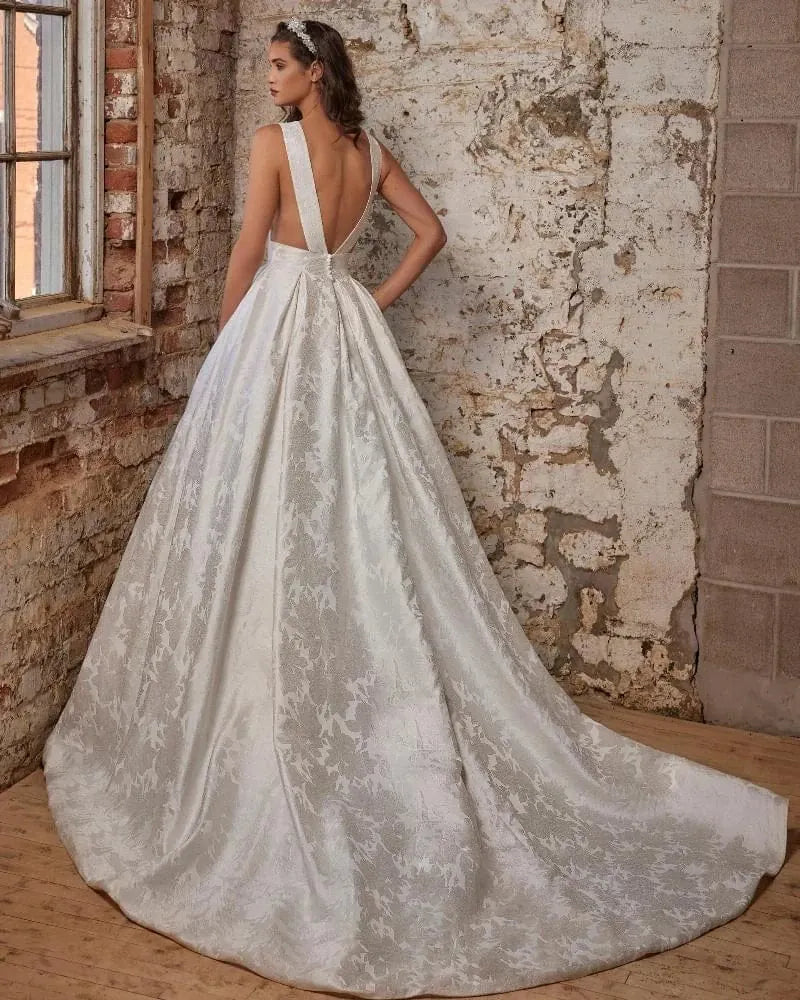 A woman modeling a white bridal gown from the back in a rustic room with exposed brick and plaster walls. The dress has a deep V back, thin straps, a fitted waist, and a full skirt with a subtle floral jacquard pattern and a long train.