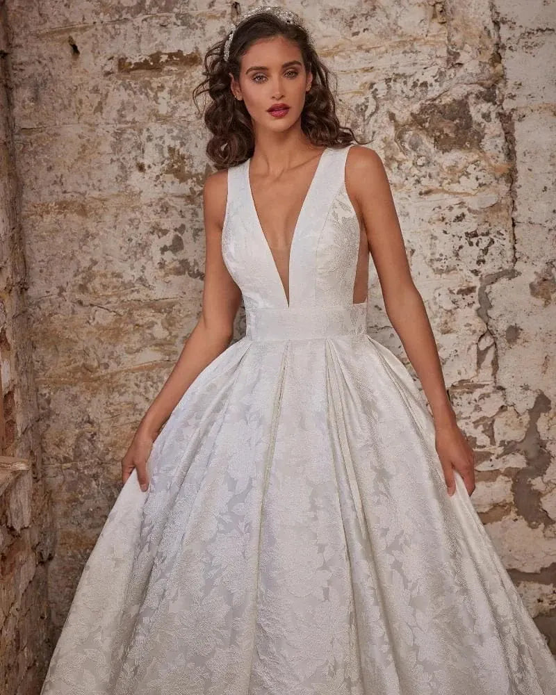 A woman wearing a white ballgown wedding dress with a deep V-neckline, lace bodice, and full textured skirt, posing against a rustic stone wall.