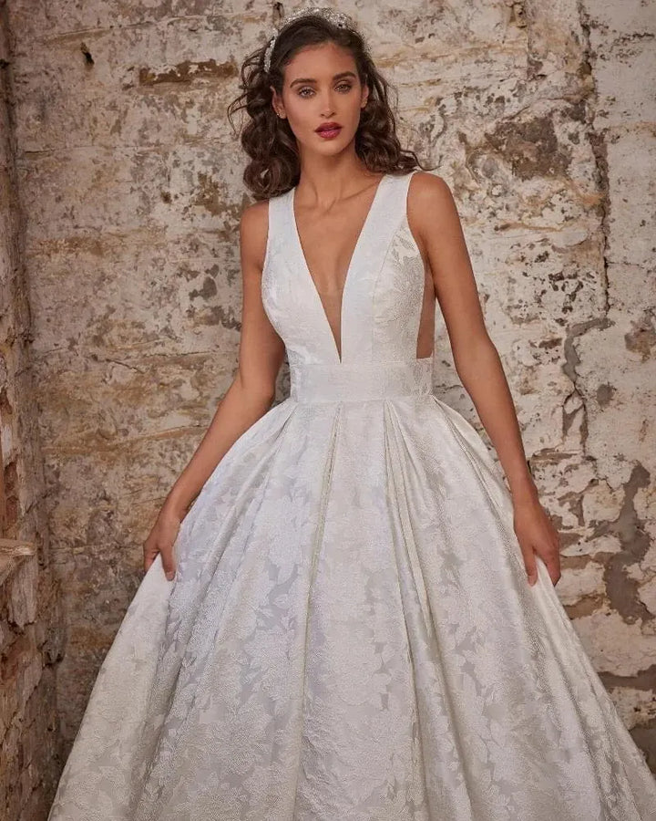 A woman wearing a white ballgown wedding dress with a deep V-neckline, lace bodice, and full textured skirt, posing against a rustic stone wall.