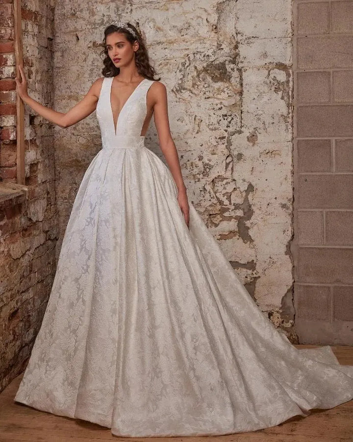 A woman models a white ballgown wedding dress with a deep V-neckline, sheer lace overlay, and a full skirt, posed against a rustic brick and plaster wall.
