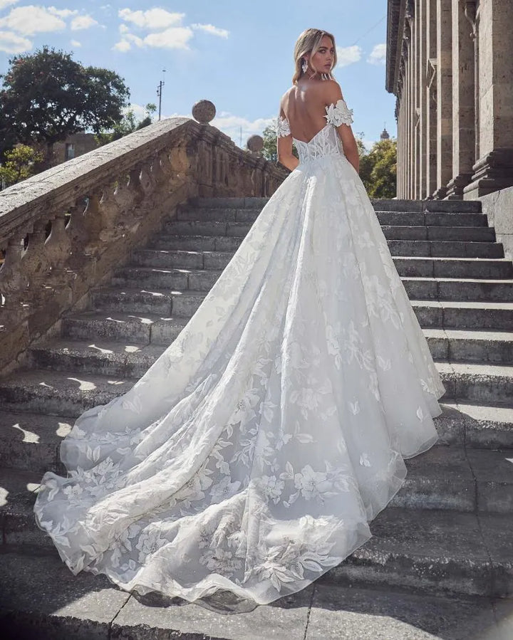 A woman wearing a voluminous white lace wedding gown with an off-the-shoulder neckline and a long train, standing on outdoor stone stairs in front of a building under a blue sky.