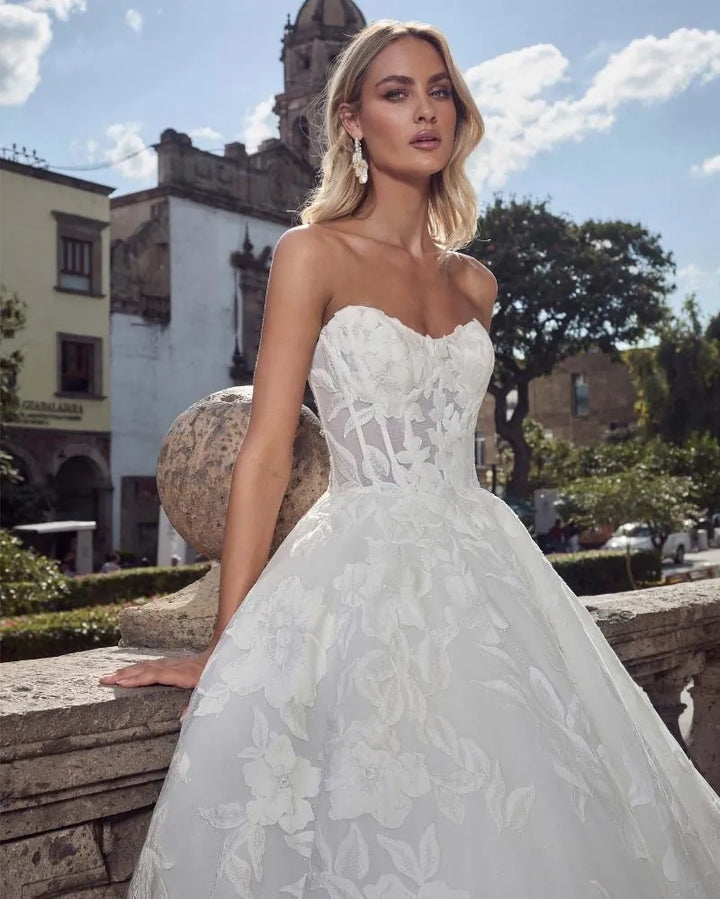 A blonde model wearing a strapless white lace wedding gown with a sweetheart neckline and a full ball gown skirt, posing outdoors on a stone balcony with a historic building and blue sky in the background.