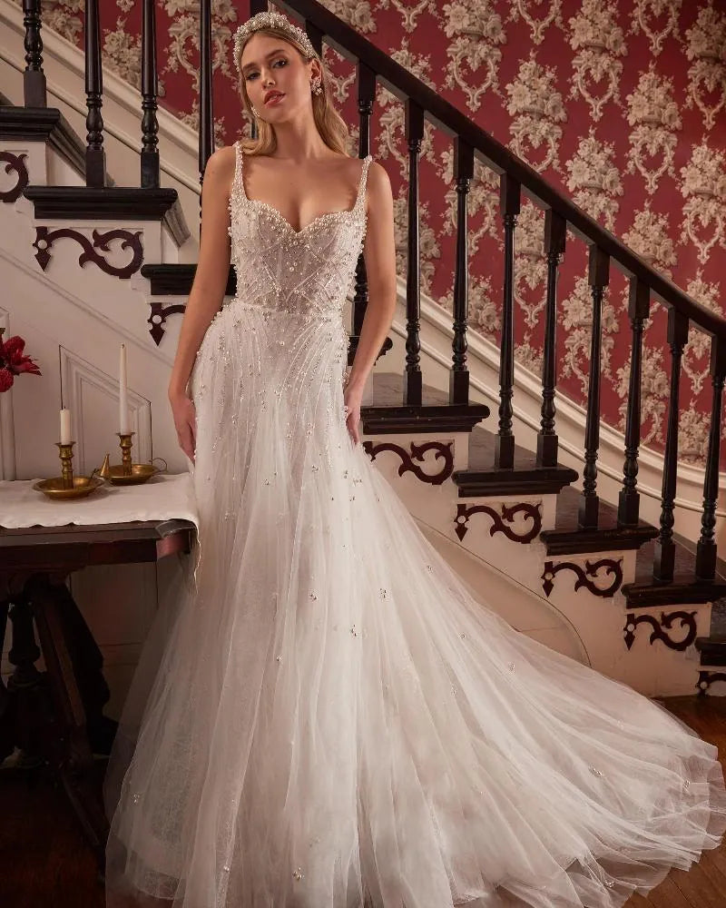 A woman wearing a white wedding gown with a sweetheart neckline, beaded bodice, and a long tulle train, standing in front of an ornate staircase with red patterned wallpaper.