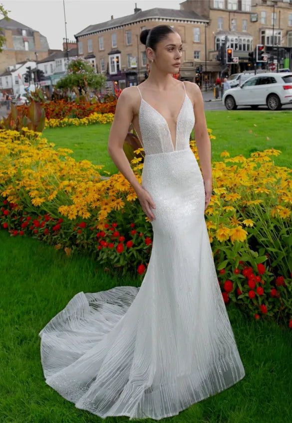 A woman wearing a white sleeveless mermaid-style wedding gown with thin straps, a deep V-neckline, and a pleated train, standing outdoors on grass in front of flower beds and town buildings.