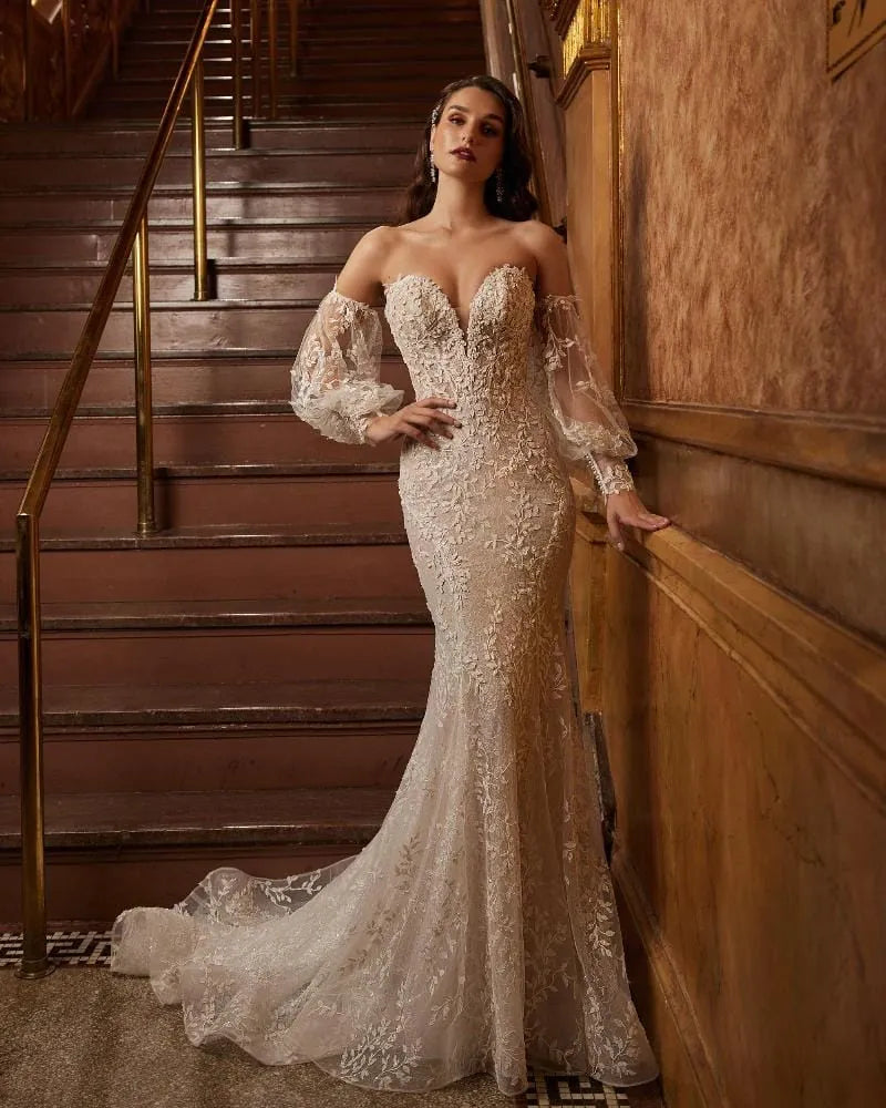A woman models an off-the-shoulder lace wedding gown with a sweetheart neckline and long sheer balloon sleeves, posed in front of an ornate wooden staircase.