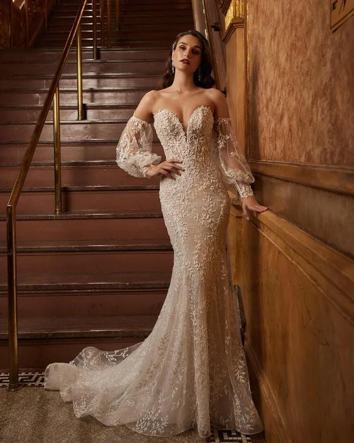 A woman models an off-the-shoulder lace wedding gown with a sweetheart neckline and long sheer balloon sleeves, posed in front of an ornate wooden staircase.