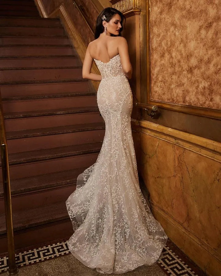 A woman wearing a strapless ivory lace mermaid gown stands on a staircase landing, photographed from the back as the dress trains on the floor.