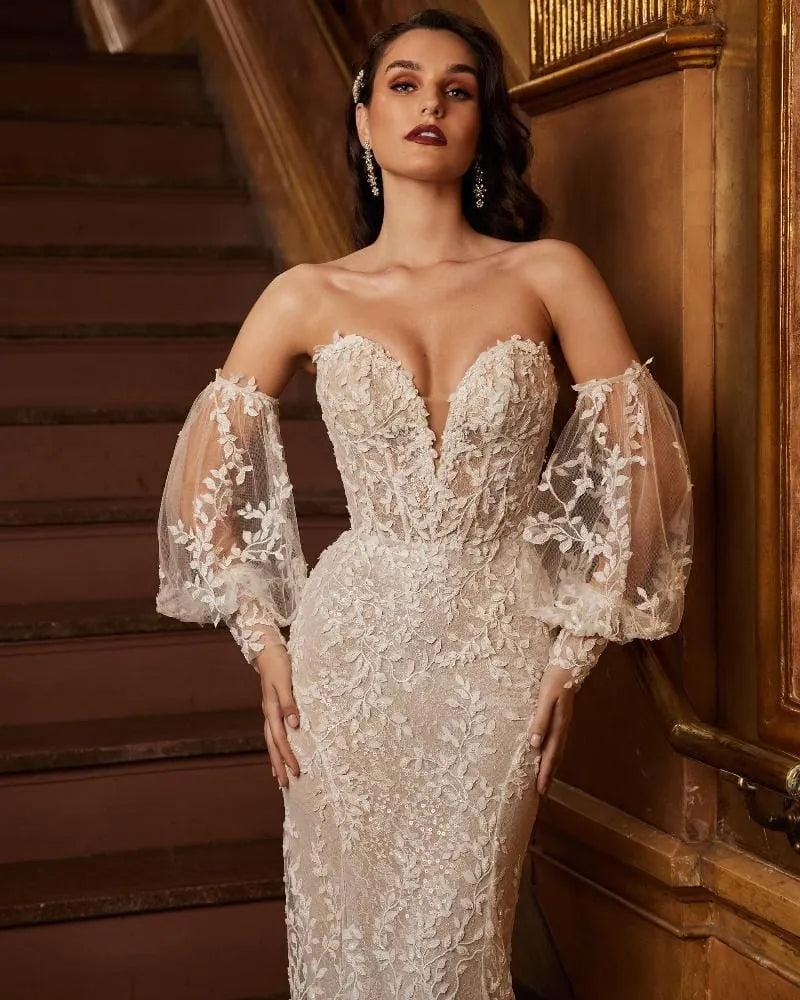 A woman models a strapless off-the-shoulder lace wedding gown with sheer balloon sleeves, standing in a warm, wood-paneled interior.
