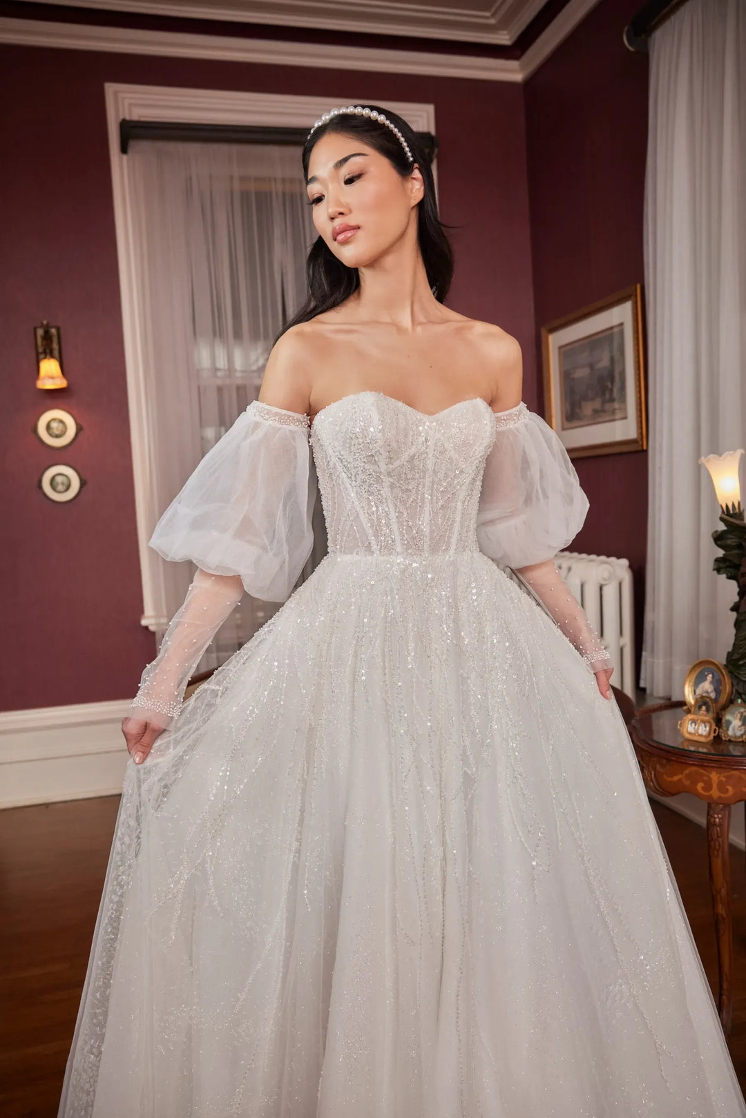 A woman wearing an off-the-shoulder ball gown wedding dress with a sweetheart neckline, fitted lace bodice, and voluminous tulle skirt, standing in a dimly lit room with burgundy walls.