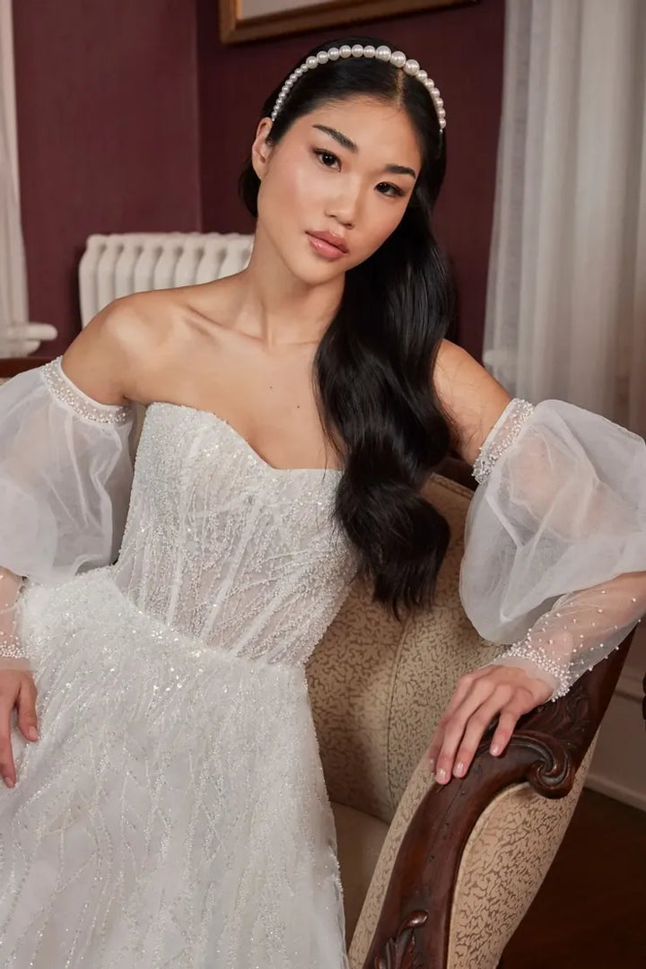 A woman wearing a white off-the-shoulder wedding dress with sheer puff sleeves and a pearl headband, seated in a vintage upholstered armchair in an indoor setting.