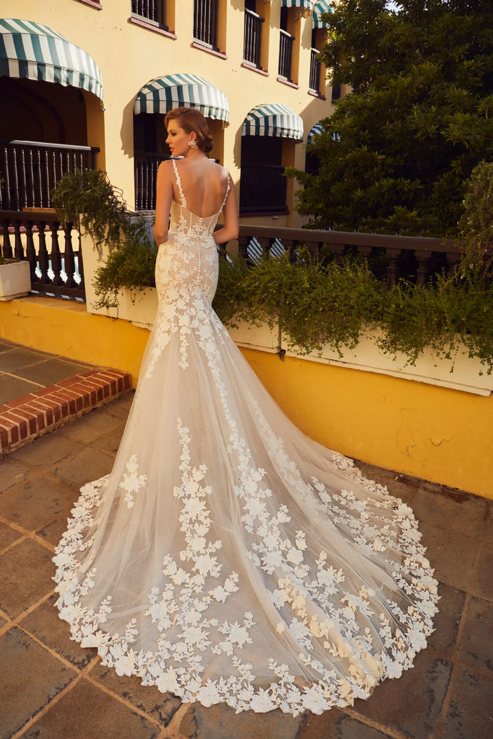 A woman wearing a fitted ivory lace wedding gown with a dramatic train, photographed outdoors on a tiled patio in front of a yellow building with striped awnings.