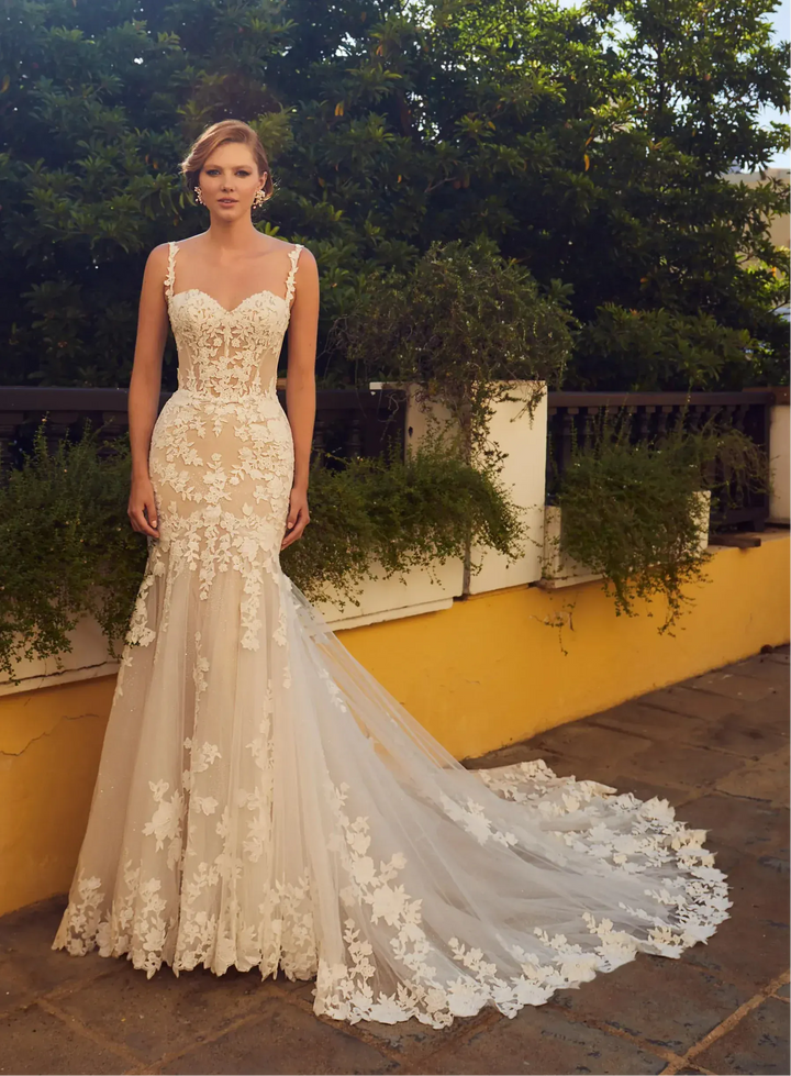 A woman wearing an elegant ivory lace mermaid wedding gown with a sweetheart neckline, thin straps, and a long sheer tulle train with floral lace appliqué, standing outdoors against a yellow wall and green foliage.