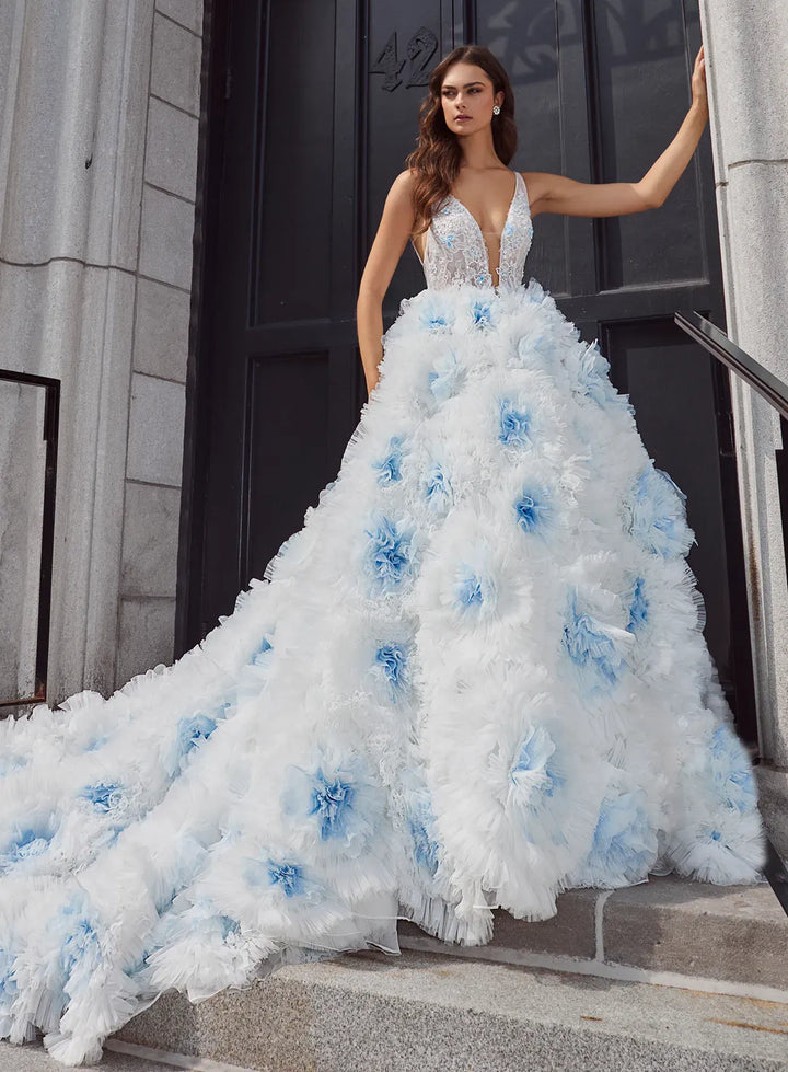 A model wears a dramatic white ball gown with light blue floral applique and a sheer lace bodice, standing on stone steps in front of a dark doorway.