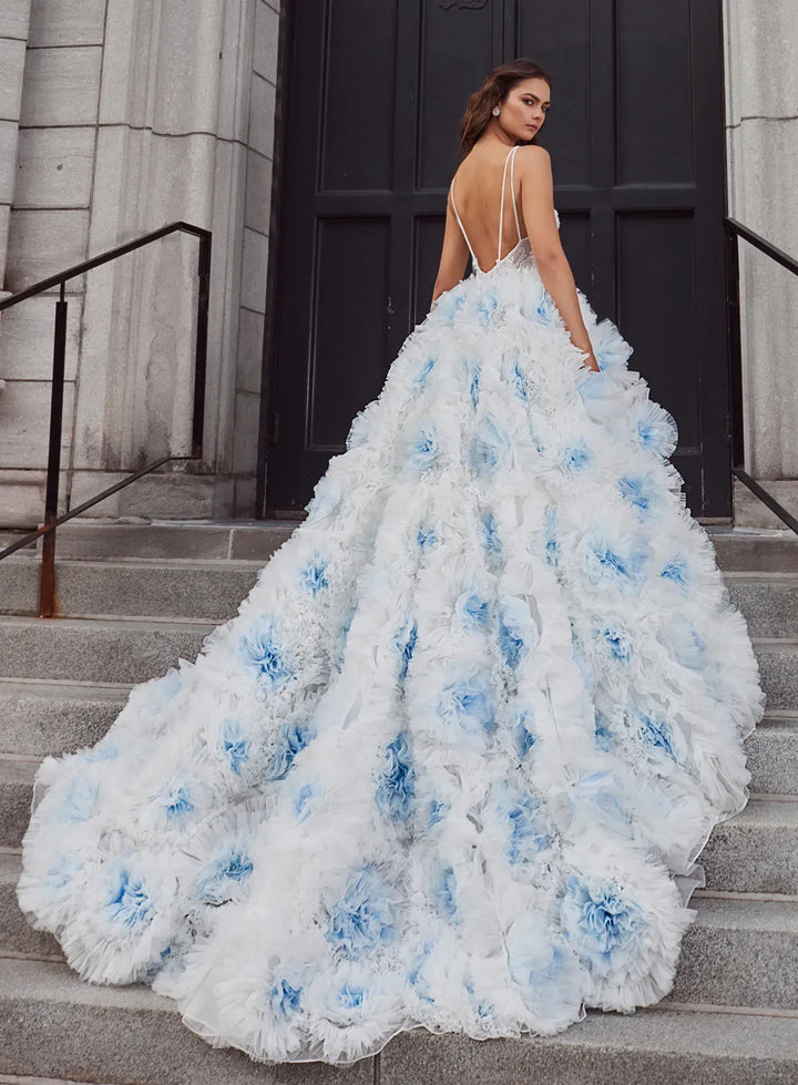 A woman stands on stone steps wearing a voluminous white ball gown with light blue floral rosettes and a backless open back, photographed outdoors in front of a dark doorway.