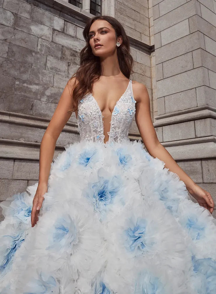 A woman models a voluminous white ball gown with light blue floral accents and a deep V-neckline, posed against a gray stone building.